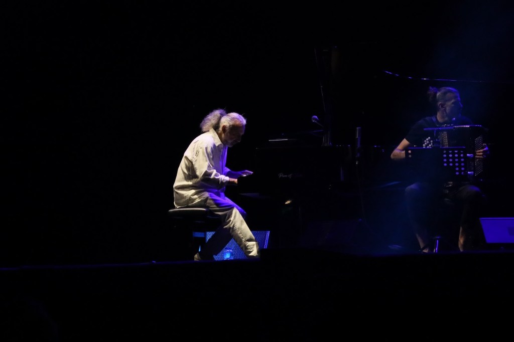 A musician playing the piano on stage with a spotlight, accompanied by another musician on an accordion.