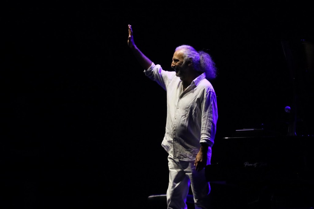 A performer on stage waves to the audience, illuminated by dramatic purple lighting against a dark background.