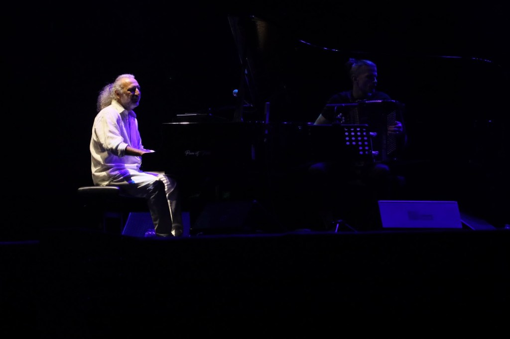 A musician playing the piano on stage while another musician plays the accordion in low light with a dark background.