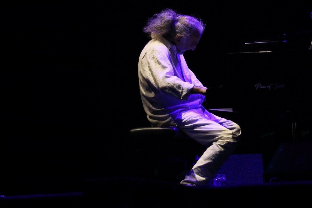 A musician playing the piano on stage under dim lighting, wearing a white outfit and focused on the keys.