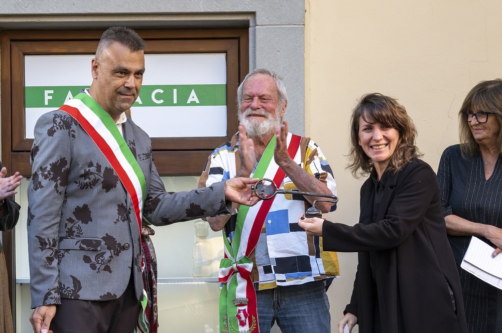 Cerimonia di consegna delle 'Chiavi della città' con un uomo in giacca con motivi floreali e due persone che applaudono, mentre una donna porge un premio.