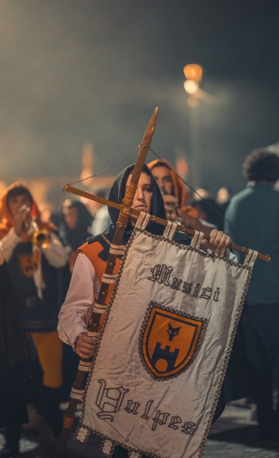 Un figurante in costume medievale tiene un banner con la scritta 'Musici Vulpes' durante le Giornate Medioevali di Poggio di Otricoli, circondato da altre persone in festa.