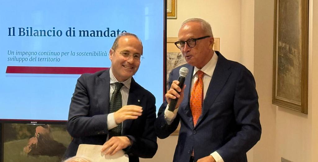 Due uomini sorridenti durante un evento, uno parla al microfono mentre l'altro lo ascolta, con uno schermo sullo sfondo che mostra il titolo 'Il Bilancio di mandate'.