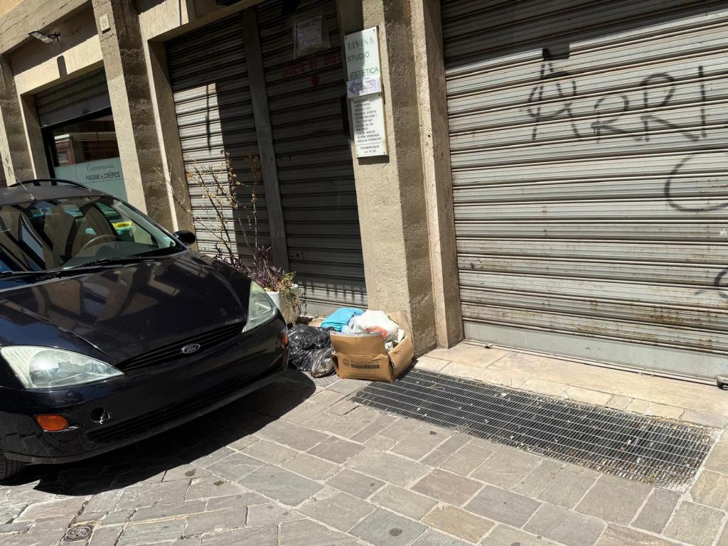 Una scena di degrado urbano a Terni, con un'auto parcheggiata accanto a un marciapiede sporco e sacchetti di immondizia abbandonati.