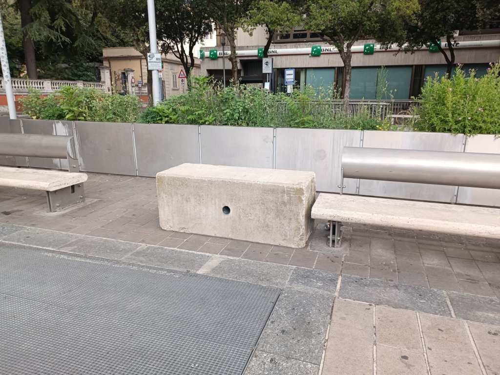 Una panchina in travertino con una seduta in cemento, circondata da un'area verde e da pannelli metallici. Sullo sfondo si intravedono alberi e edifici.