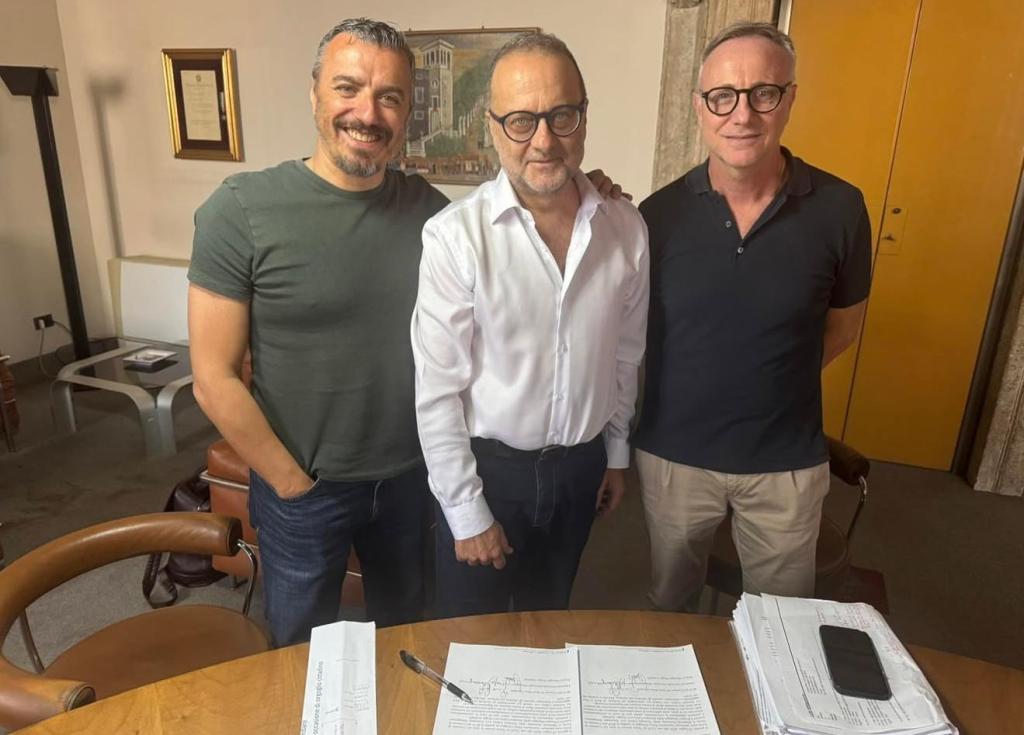 Tre uomini sorridenti posano insieme davanti a un tavolo con documenti e una penna, in un ambiente interno con decorazioni classiche.