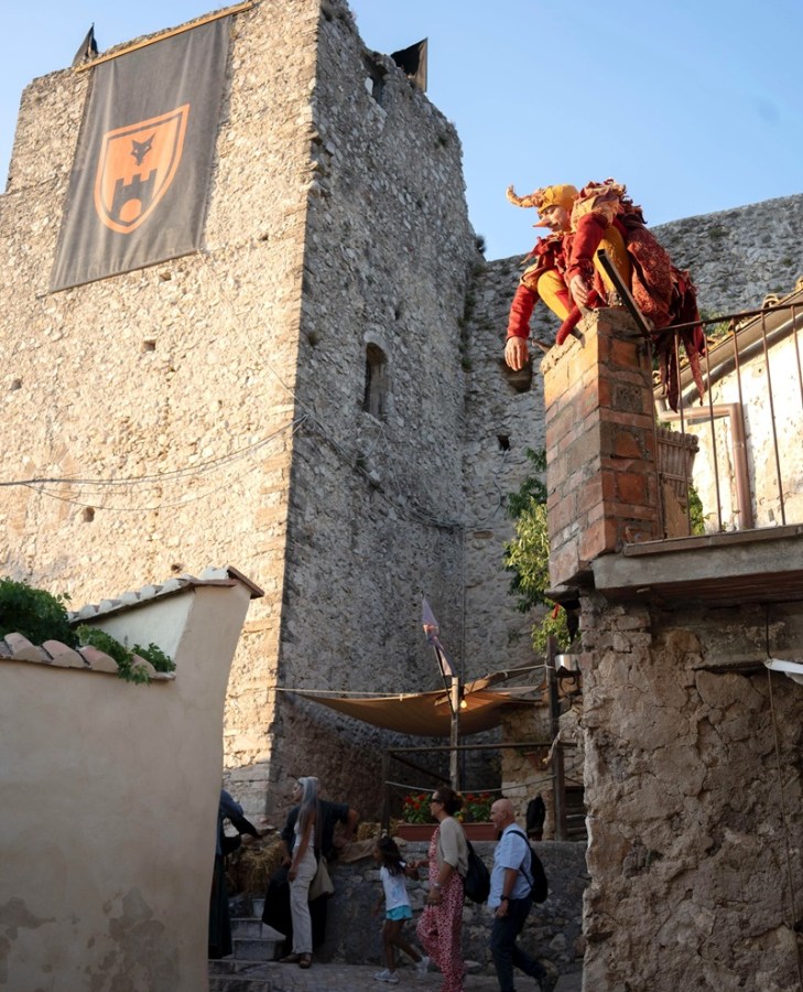 A performer in a medieval costume sitting on a ledge near an ancient stone tower, with visitors walking below in a picturesque setting of a historic village during a festival.