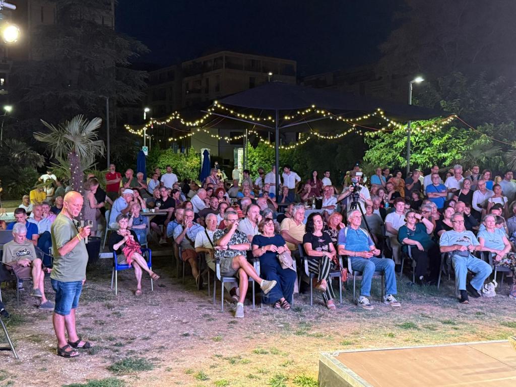Un pubblico numeroso partecipa a un evento all'aperto di notte, con una persona che parla al microfono sul palco.