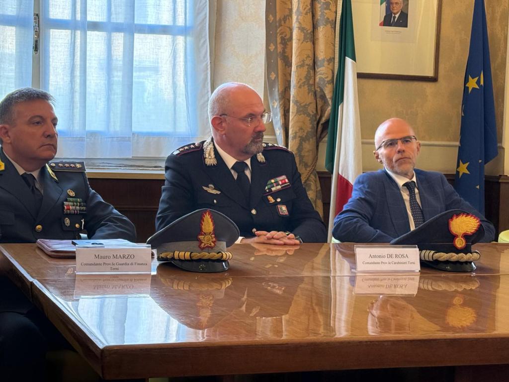 Three men seated at a conference table in a formal setting, with flags in the background. The first man, dressed in a military uniform, wears various insignia. The second man, also in a uniform, has a serious expression. The third man, in a suit and glasses, appears thoughtful.