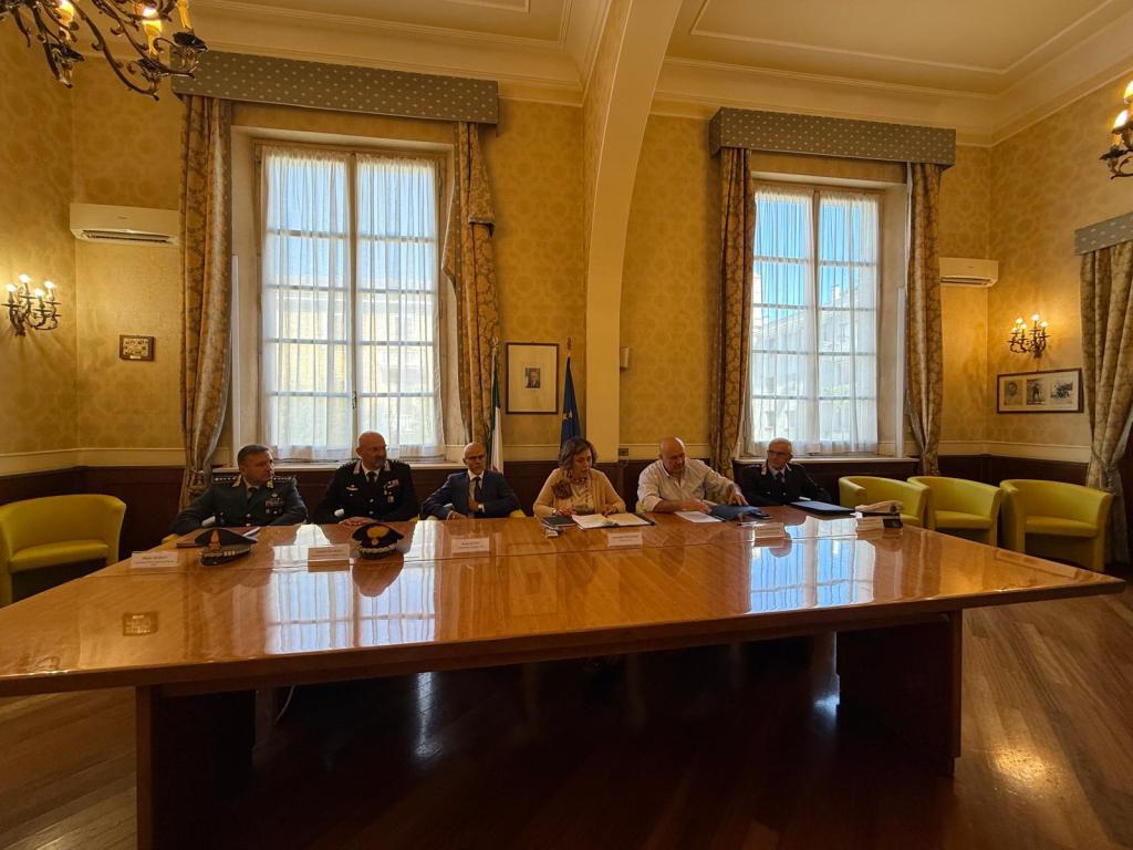A group of officials seated around a large table in a well-lit room with yellow walls and elegant decor. The scene includes five individuals, some in uniforms, engaging in a discussion with documents and a flag in the background.