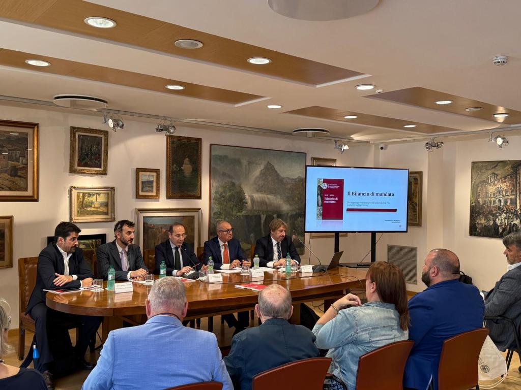 A group of five men seated at a table during a presentation, with a large screen displaying the title 'Il Bilancio di mandato' in the background. The room features various paintings on the walls and an audience listening attentively.