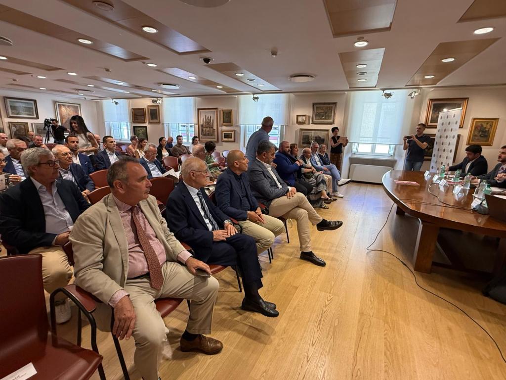 A conference or meeting room filled with people seated in rows, facing a table with speakers. The room features artwork on the walls and windows with natural light coming through.