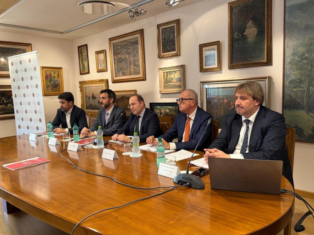 A group of five individuals seated at a wooden table during a conference, with water bottles and documents in front of them, surrounded by framed art on the walls.