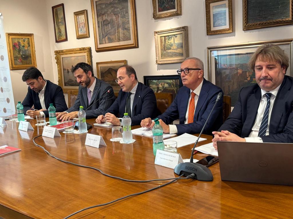 Panel discussion featuring five seated individuals at a table with water bottles and papers, surrounded by framed artworks on the walls.