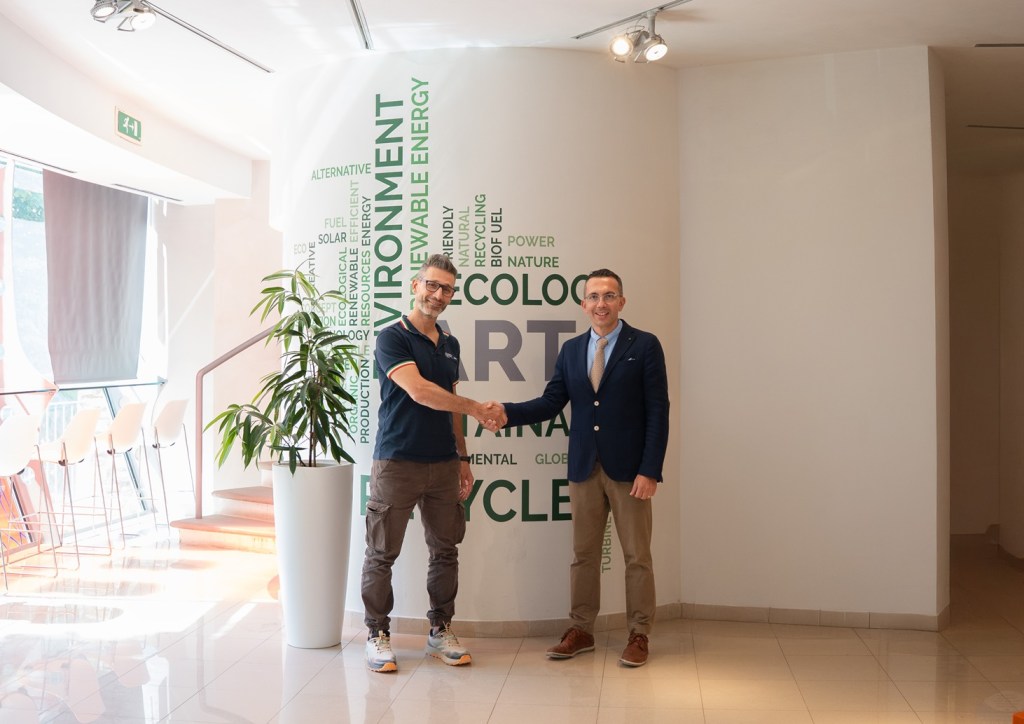 Due uomini si stringono la mano in un ambiente moderno, con pareti decorate da parole chiave relative alla sostenibilità e all'ambiente.