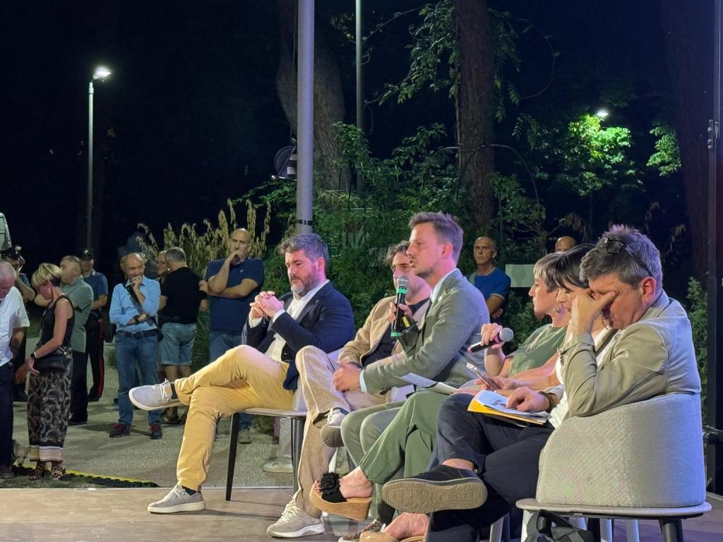 Un gruppo di relatori partecipa a un incontro pubblico, seduti su seggi, mentre il pubblico ascolta in uno spazio all'aperto di notte.