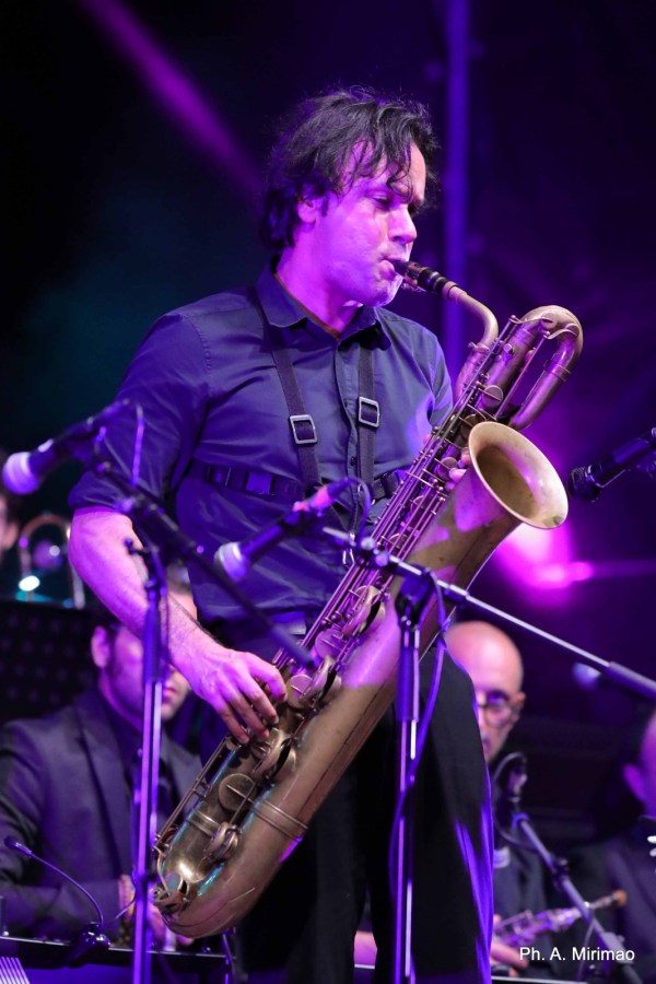 A musician playing a saxophone on stage, featuring vibrant purple lighting and an engaged audience in the background.
