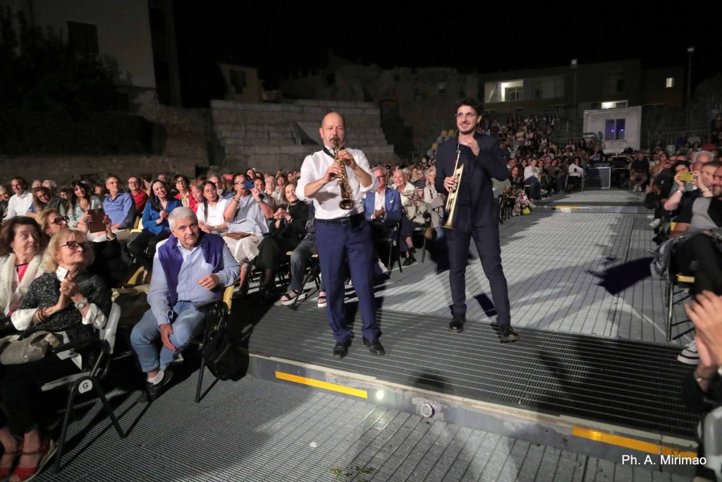 A live outdoor performance featuring two musicians, one playing the saxophone and the other holding a trumpet, in front of an audience seated in chairs at night.