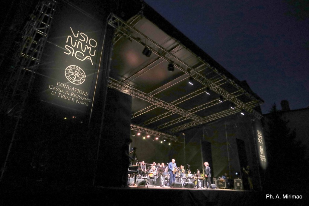 Stage view of a concert event at night, showcasing performers and a large banner with 'Visio Musica' and sponsor details.