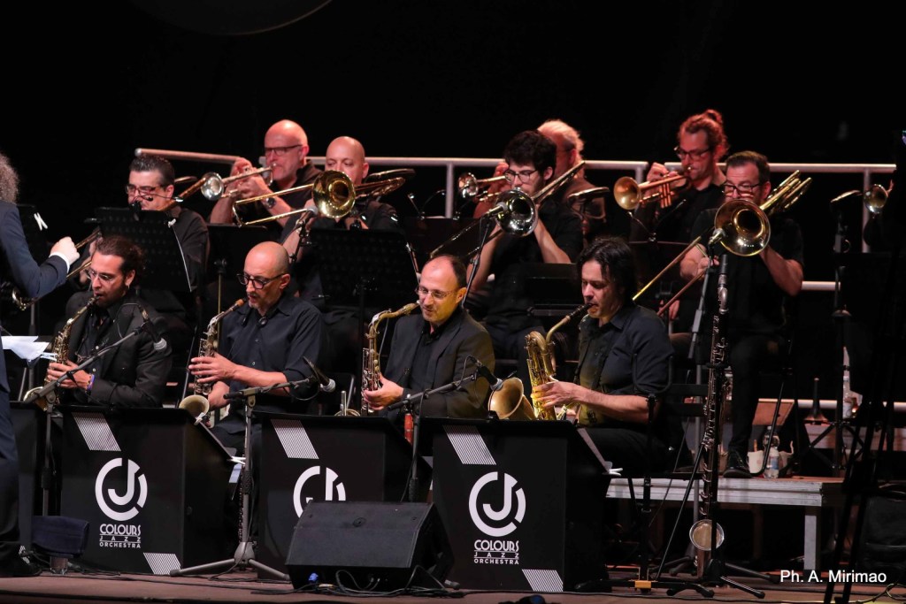 A jazz orchestra performing on stage, featuring musicians playing various brass instruments, including saxophones and trombones, dressed in black attire.