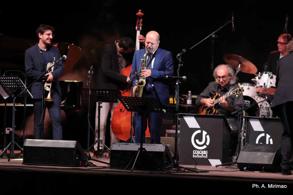 A jazz band performing live on stage with a saxophonist in a blue suit, a trumpet player, a guitarist, and a bassist, surrounded by music stands and instruments.