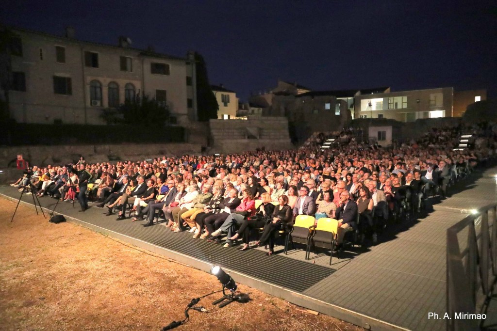 A large crowd of people seated in an outdoor theater at night, with a mix of empty and occupied seats, and buildings in the background.
