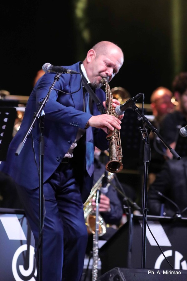 A musician performing passionately on stage while playing a saxophone, dressed in a formal blue suit, with a band in the background.