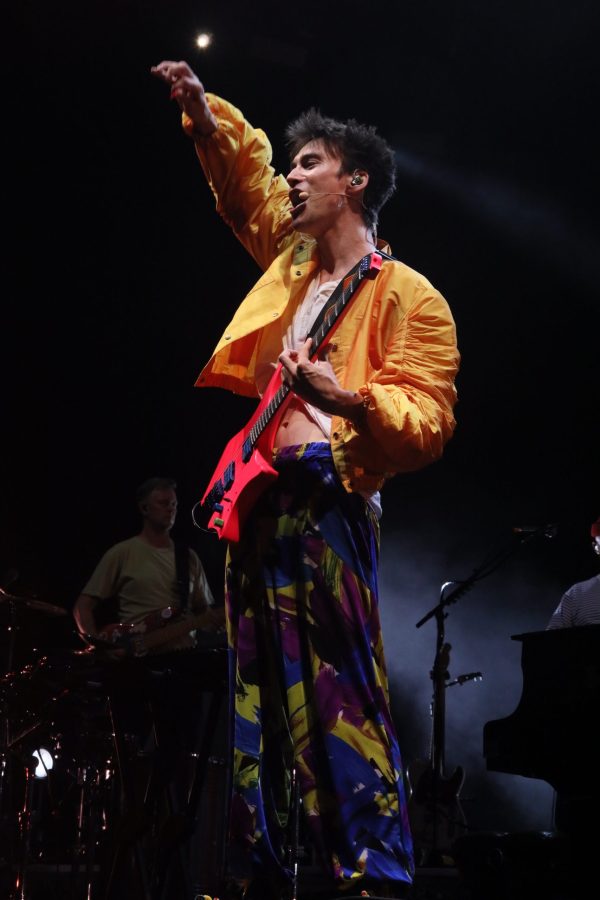 Un artista sul palco durante un concerto, indossando un giubbotto giallo e pantaloni colorati, mentre suona una chitarra elettrica rossa.