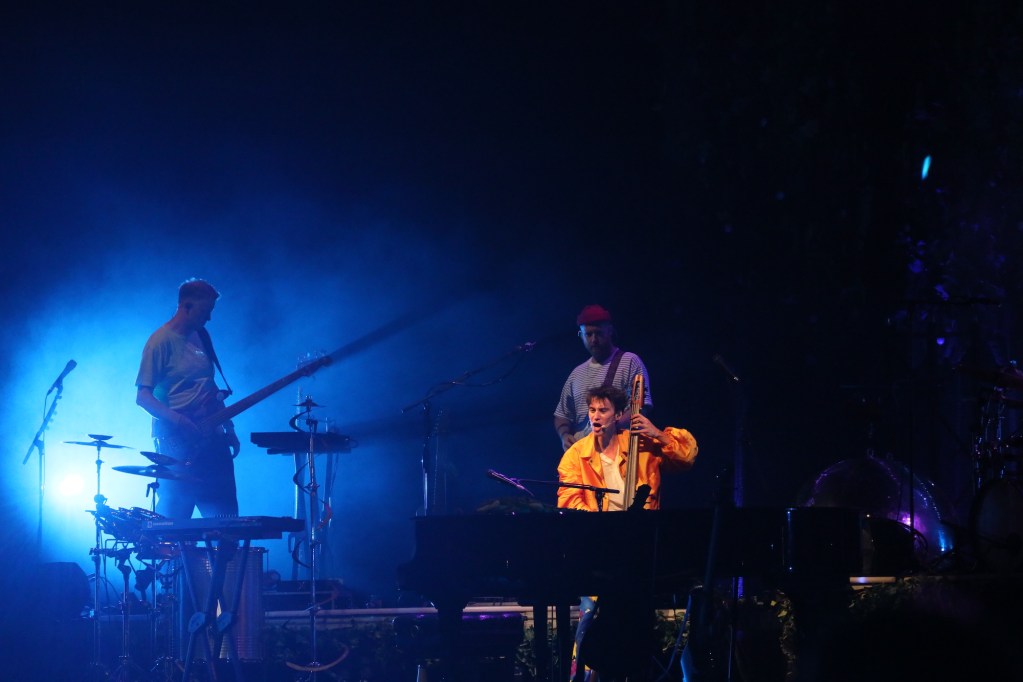 Un artista suona il pianoforte dal vivo, circondato da luce blu e luci di scena, mentre un musicista suona la chitarra sullo sfondo.