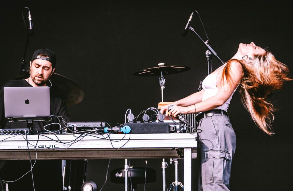 Un'artista si esibisce sul palco mentre balla e canta, accanto a un produttore che utilizza un laptop e strumenti musicali.