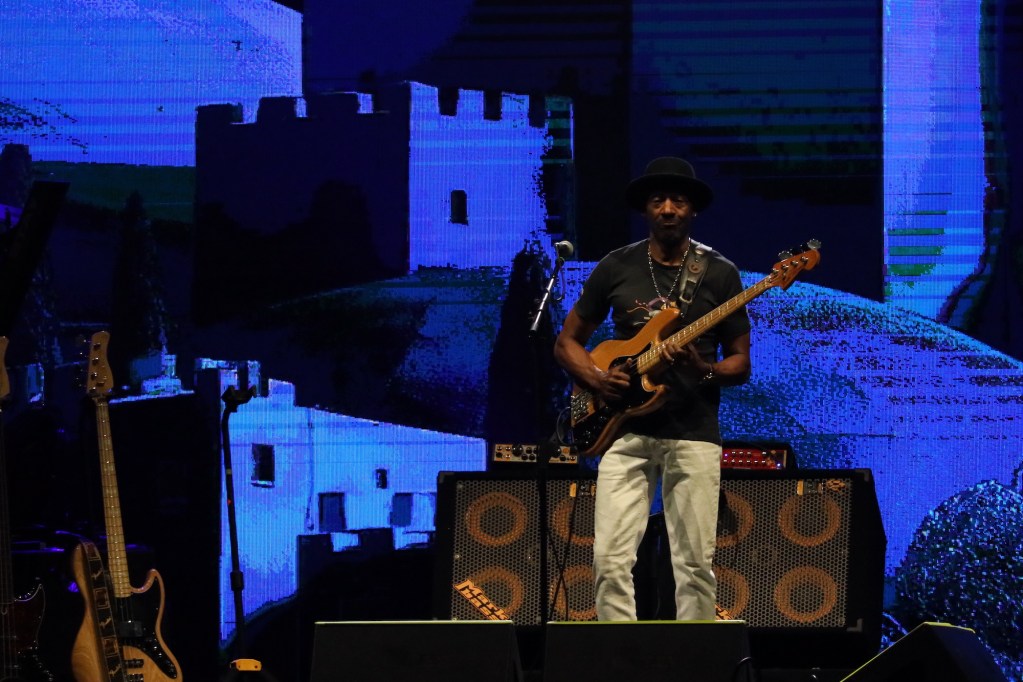 Marcus Miller suona il basso sul palco con un grande schermo sullo sfondo che mostra un paesaggio astratto.