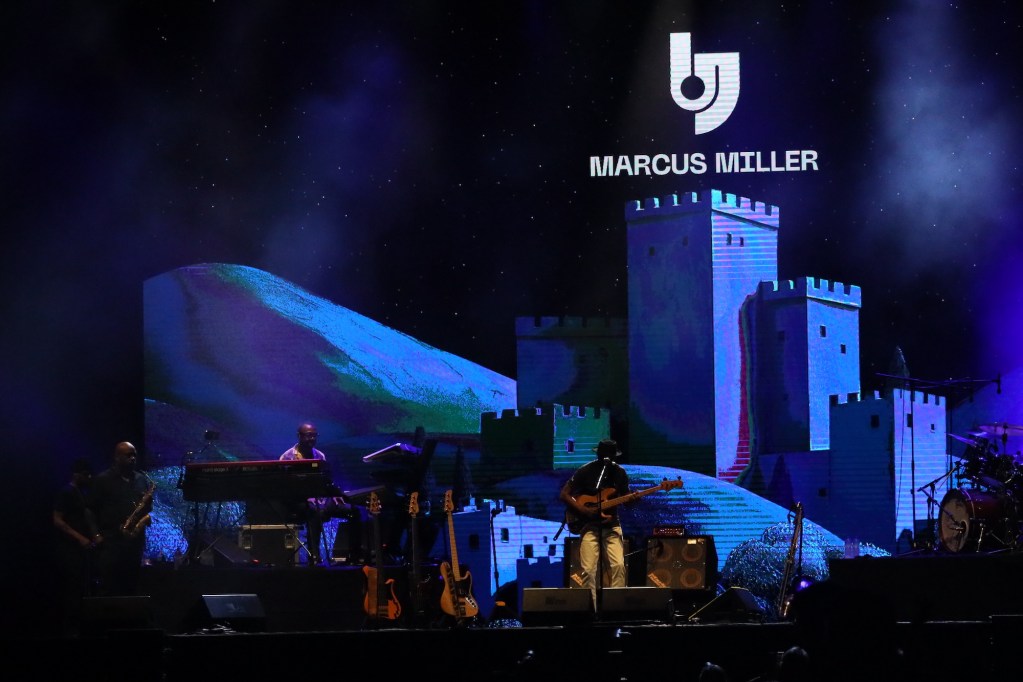 Marcus Miller performs on stage at the Arena Santa Giuliana, with a colorful backdrop featuring a castle and musical instruments in front.