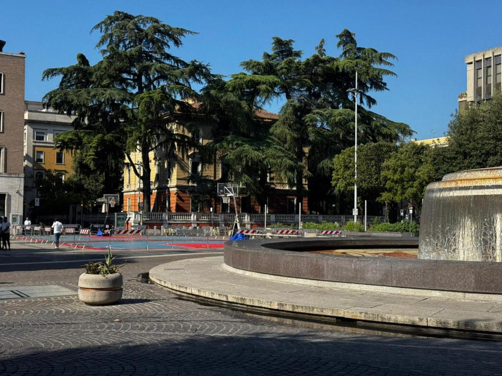 A sunny urban area featuring a large fountain and several tall trees. In the background, there are buildings, including a yellow structure. A basketball court is visible and surrounded by construction barriers.