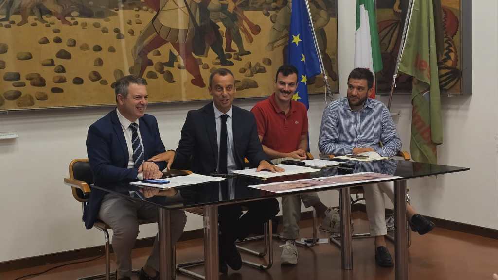 Quattro uomini sorridenti seduti attorno a un tavolo in una sala con bandiere e un grande dipinto sullo sfondo, durante un incontro dedicato all'Agosto Corcianese.