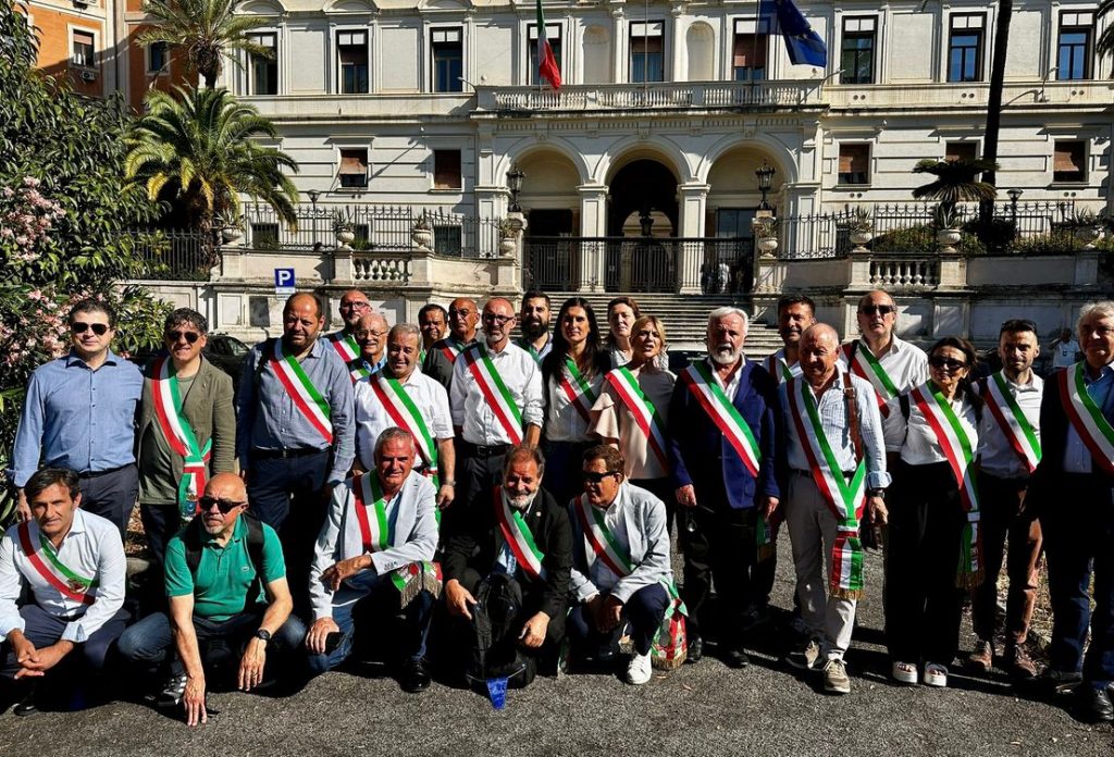 Group of individuals wearing sashes displaying the colors of the Italian flag, posing in front of a historic building with palm trees and flags.