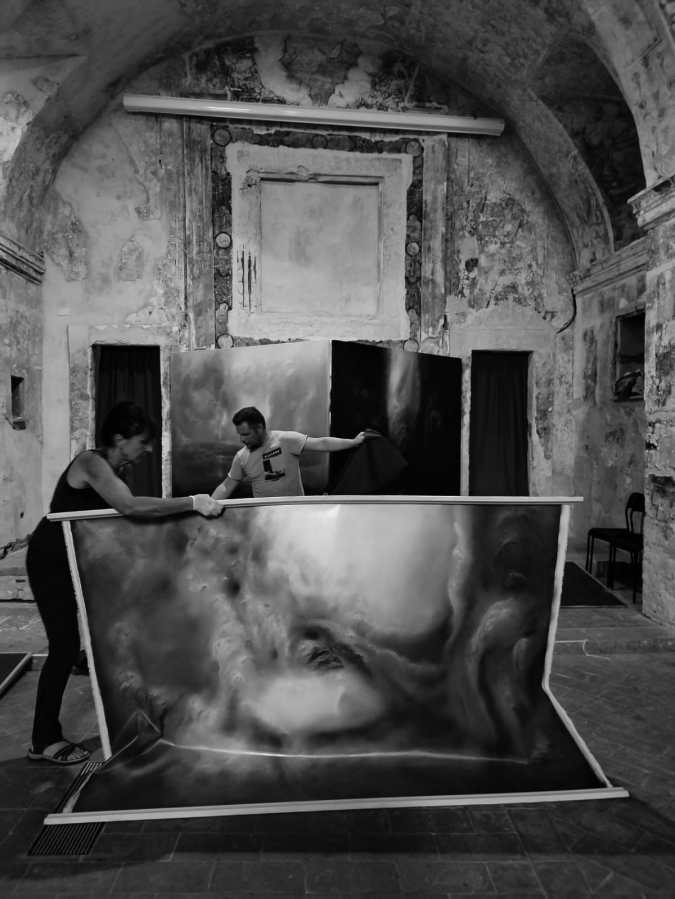 Due persone stanno allestendo grandi tele d'arte in un'ambientazione rustica, con pareti in pietra e decorazioni storiche, all'interno di una chiesa.