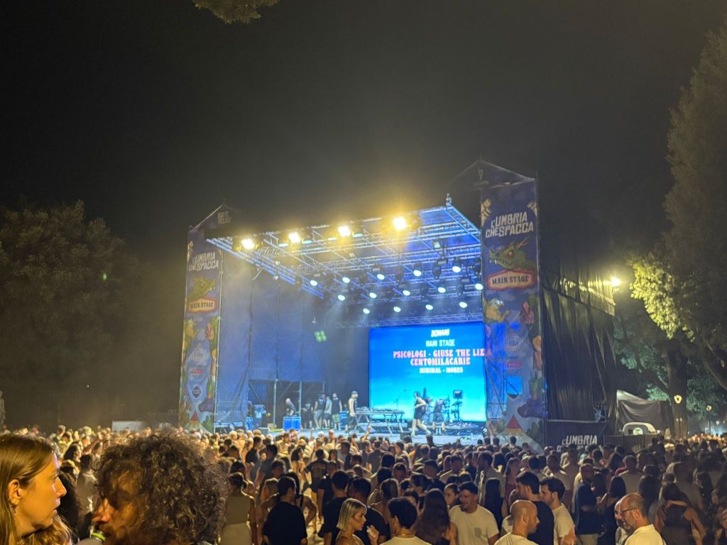 Concerto serale al festival Umbria Che Spacca, con una folla numerosa di persone davanti al palco illuminato, mentre un artista si prepara per la performance.