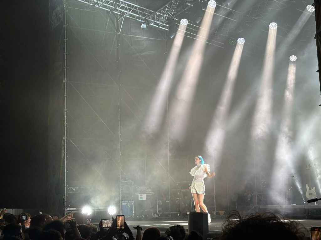 Una performer sul palco durante un concerto, circondata da luci intense e fumi, mentre il pubblico rivolge le sue attenzioni verso di lei.