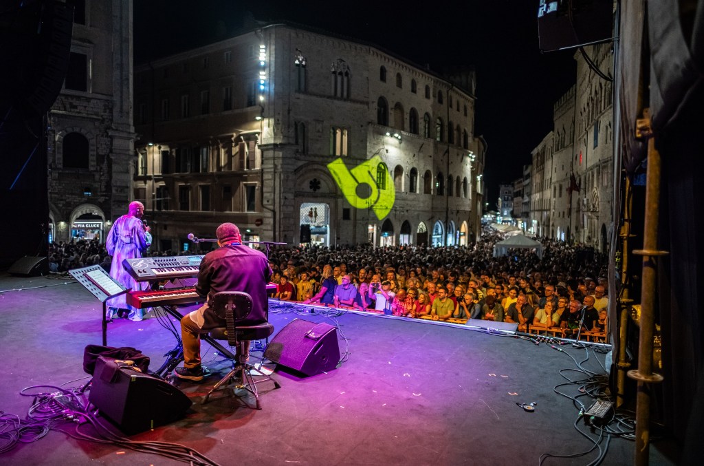 A vibrant outdoor concert scene at night, featuring a musician on stage performing for a large crowd, with illuminated historic buildings in the background.