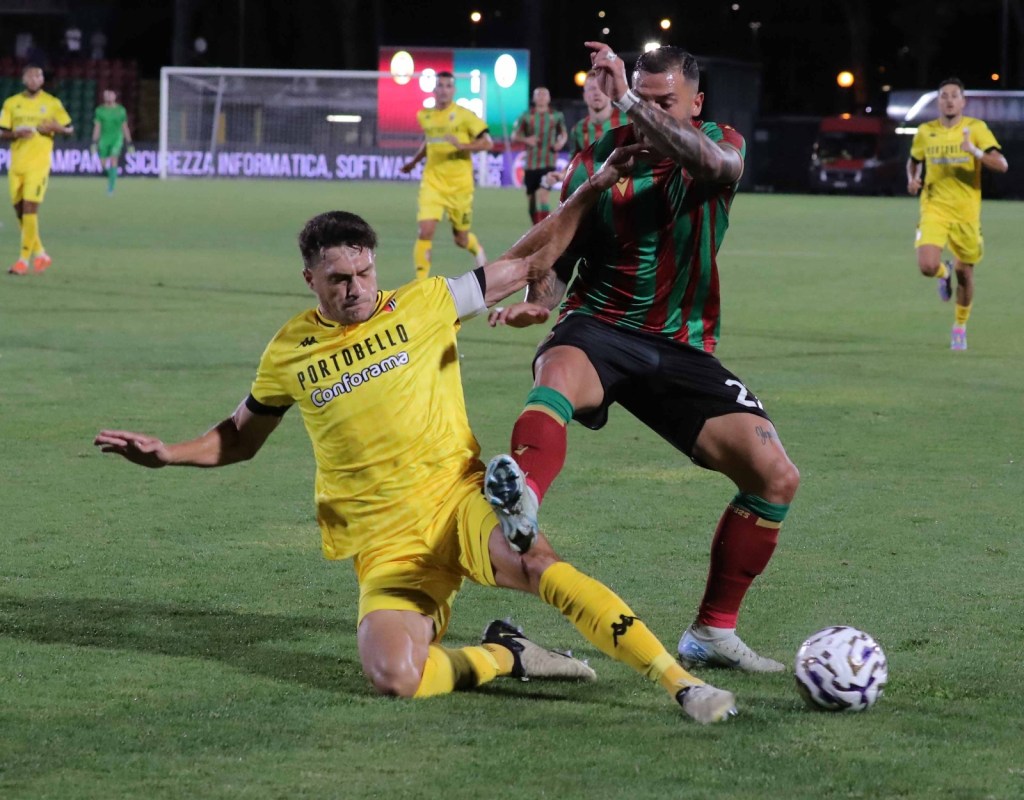 A soccer match scene featuring two players in action, with one player in a yellow jersey attempting to tackle the other in a striped green and red jersey. The background shows other players and a scoreboard.