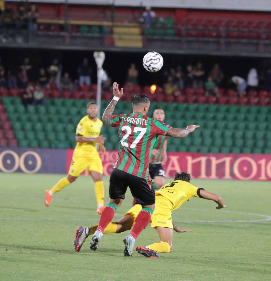 A soccer player in a red and green striped jersey, wearing the number 27, prepares to pass the ball while another player in a yellow jersey falls to the ground. The background includes spectators and a green seating area.