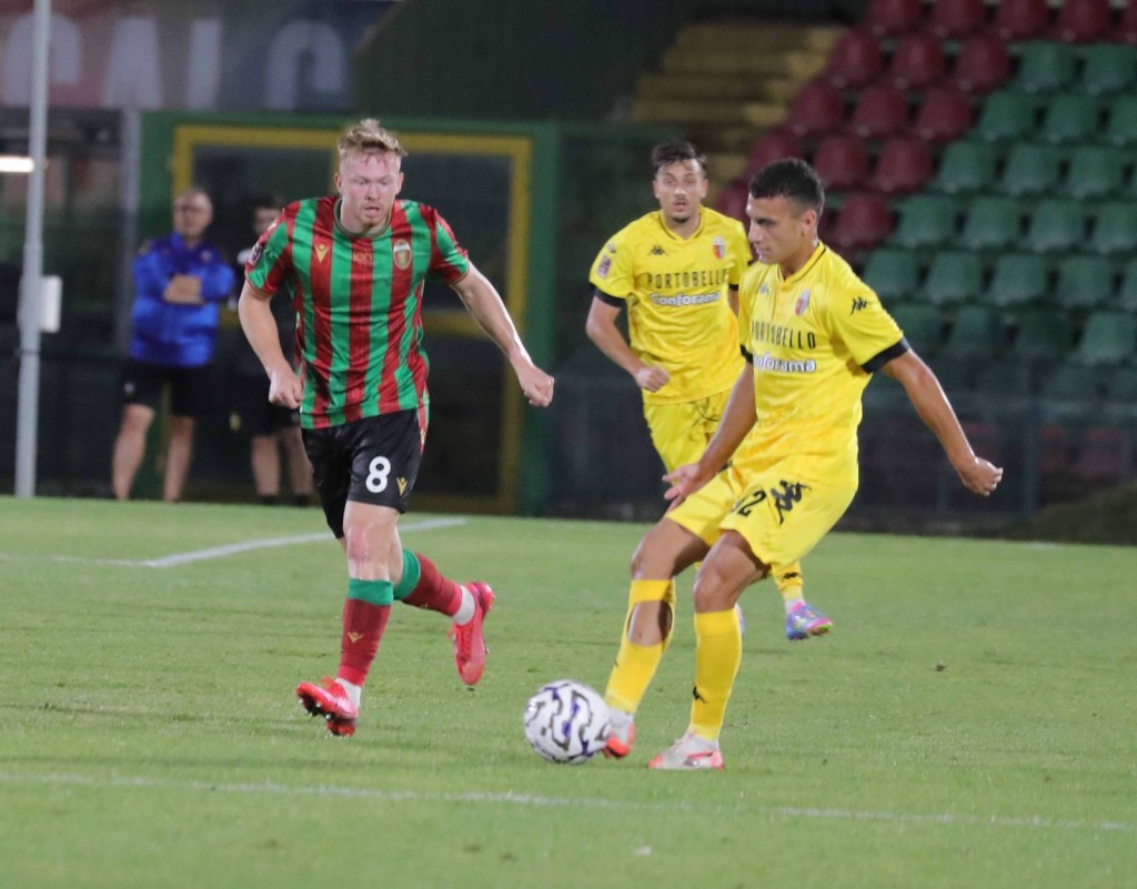 Un giocatore in maglia verde e rossa corre mentre un avversario in maglia gialla si prepara a colpire il pallone durante una partita di calcio.