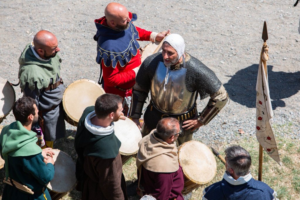 Un gruppo di persone in abiti medievali si riunisce attorno a tamburi, mentre un cavaliere in armatura discute con loro.