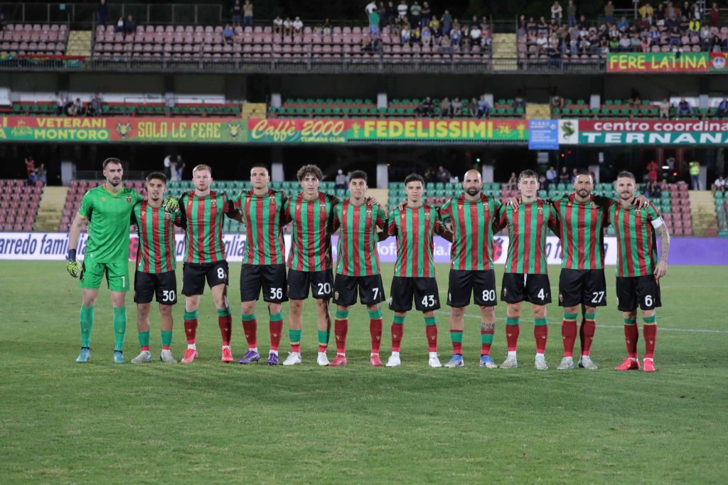 A soccer team poses for a group photo on the field, wearing green and red striped jerseys and black shorts, with an enthusiastic crowd and stadium seating in the background.