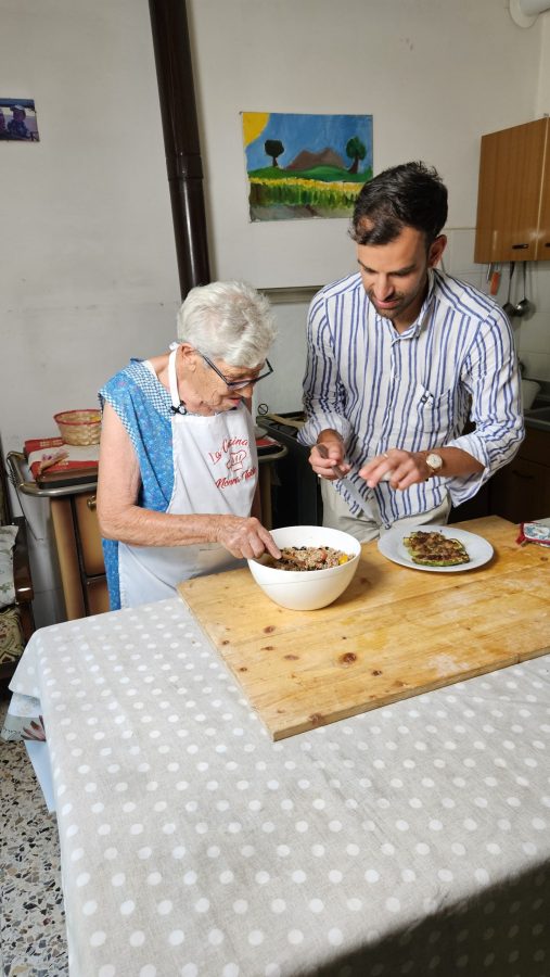 Una donna anziana e un giovane uomo stanno preparando piatti nella cucina di casa, con un tavolo di legno e una tovaglia a pois. Sul tavolo ci sono ciotole con ingredienti e un piatto servito. Alle pareti ci sono opere d'arte.