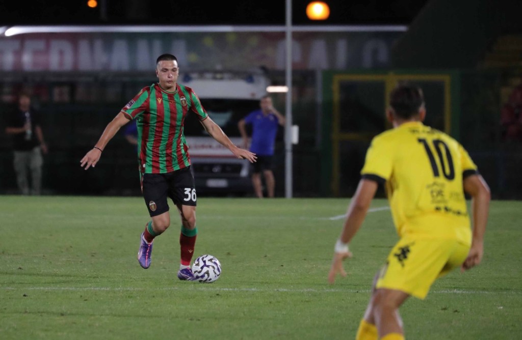 A football player in a green and red striped jersey with the number 36 is dribbling the ball on the field, while another player in a yellow jersey with the number 10 is positioned nearby.