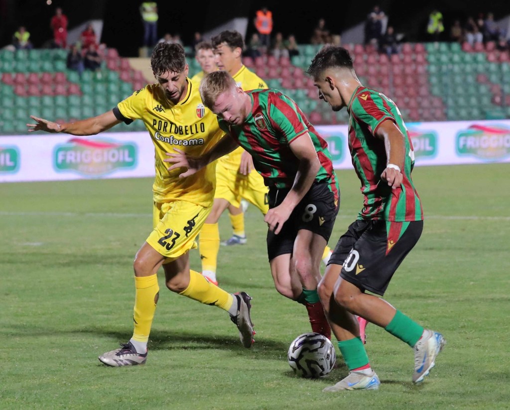 Two soccer players in jerseys, one yellow and one red-green, are engaged in a competitive play with the ball on the field.