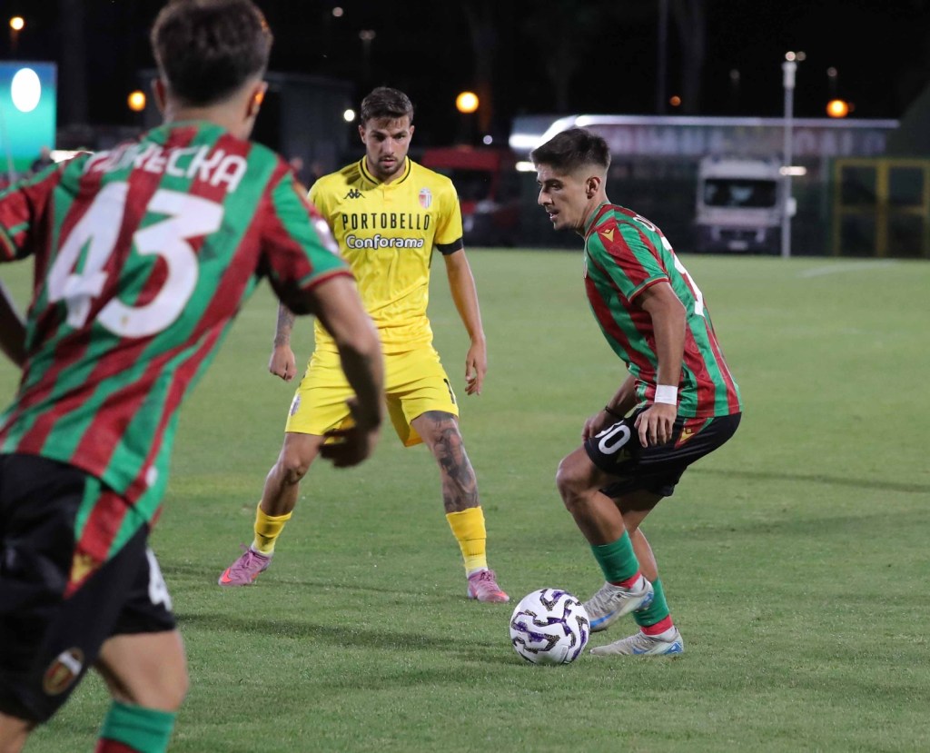 Two football players in action on the field, one wearing a yellow kit and the other in a red and green striped jersey with the number 10, while a third player in a similar striped kit approaches from the left.
