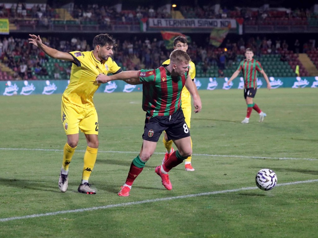 A soccer match scene showing two players competing for the ball on a grassy field, with a crowd visible in the background.