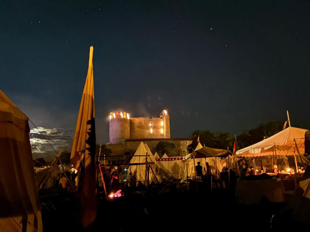 Vista notturna di un castello illuminato con tende di rievocazione storica in primo piano.
